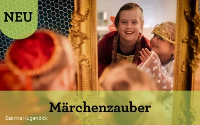 Zwei Mädchen in Prinzessinnenkleidern schauen lachend in einen golden umrandeten Spiegel